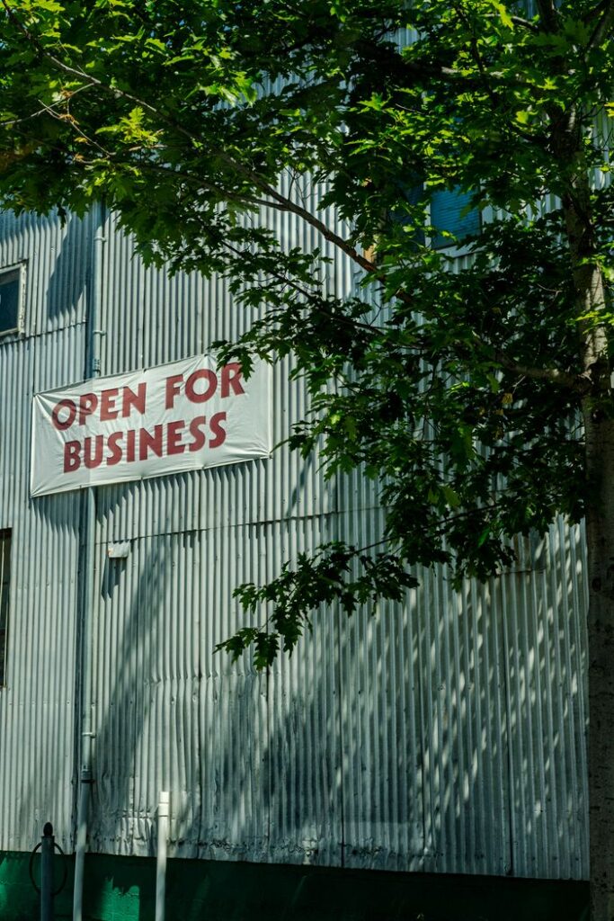 Industrial building facade with 'Open for Business' sign shaded by tree branches.